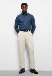 Marineblaues Hemd mit Knopfleiste und kleinem weißem Punktmuster, kombiniert mit beigen Hosen und dunklen Schnürschuhen. Schlanke Passform.