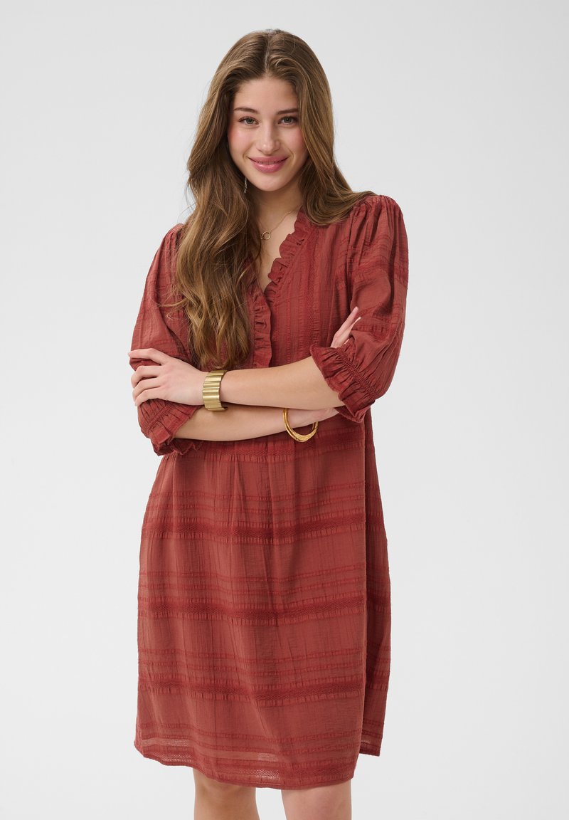 Jeune femme aux longs cheveux ondulés porte une robe couleur rouille, longueur genoux, avec des manches trois-quarts et des bracelets en or, souriant les bras croisés.