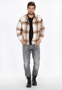 Karo-Jacke in Beige, Braun und Weiß, mit Knopfverschluss und gerippten Bündchen. Getragen über einem schwarzen Hemd mit grauen Jeans und schwarzen Stiefeln.