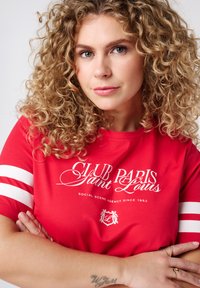 T-shirt rouge avec un texte blanc "CLUB PARIS Saint Louis," présentant des rayures blanches sur les manches et une texture douce en coton.