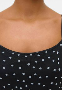 Débardeur noir à pois blancs, avec un col rond et des bretelles fines, fabriqué en tissu lisse avec une légère brillance.