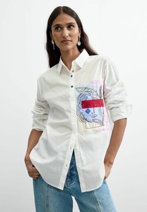 Frau trägt ein weißes Hemd mit Knöpfen und einem bunten abstrakten Gesichtspatch auf der Brust, kombiniert mit blauen Jeans und hängenden Ohrringen.