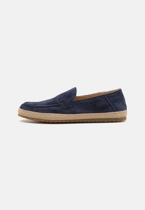 Chaussure slip-on en suede bleu marine avec une semelle en jute naturel tissée et текстurée. Présente un profil bas et des détails de couture minimalistes sur le dessus.