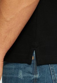 Schwarzes Polohemd aus strukturiertem Material, mit einem Schlitz am Saum und einem sichtbaren Knopfakzent, kombiniert mit blauen Jeans.