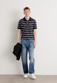 Mørk marineblå stripete polo skjorte, blå jeans, hvite sneakers, holder en svart blazer. Fokus på avslappet, tilpasset design og fargekontrast.