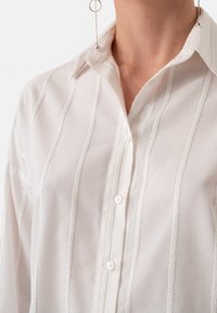 White button-up shirt with vertical silver stripes, featuring a classic collar, textured fabric, and small white buttons down the front.