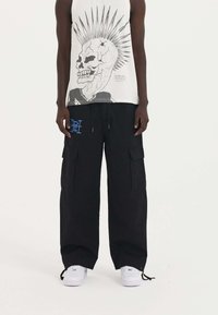 Schwarze Cargo-Hosen mit zwei Seitentaschen und blauen Logo-Details, kombiniert mit einem weißen ärmellosen Shirt mit einem Totenkopf-Graphic.