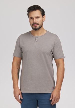 Mężczyzna z brodą, ubrany w szarą koszulkę henley z krótkim rękawem i niebieskie spodnie, stojący na jednolitym jasnym tle.