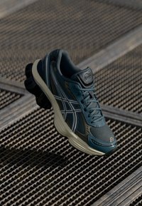 Chaussure de course pour hommes avec une tige en mesh de couleur vert foncé, ornée d'accents argentés et d'une semelle rembourrée. La semelle extérieure présente un design incurvé distinctif.