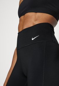 Melni augstās sporta legingi ar gludu tekstūru, ar baltu Nike logotipu un plakanšuvēm, kas nodrošina papildu izturību.