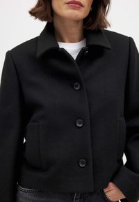 Schwarzer, kurzer Wollmantel mit Kragen, ausgestattet mit einer Knopfleiste und zwei seitlichen Taschen. Weiche Textur und minimalistisches Design.