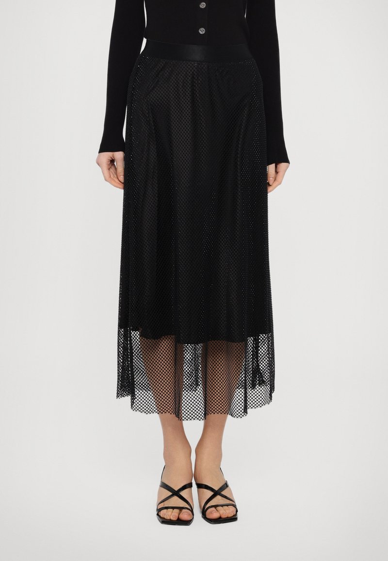 Saia midi preta com sobreposição de rede transparente e cós sólido, apresentando padrões texturizados. Combinada com sandálias de salto pretas com tiras.