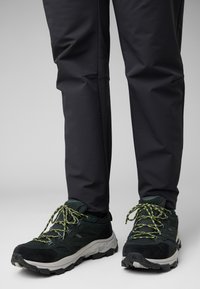 Scarpe da esterno nere con materiale testurizzato e suola spessa, caratterizzate da lacci e dettagli verde brillante, abbinate a pantaloni scuri.