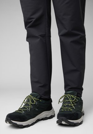 Scarpe da esterno nere con materiale testurizzato e suola spessa, caratterizzate da lacci e dettagli verde brillante, abbinate a pantaloni scuri.