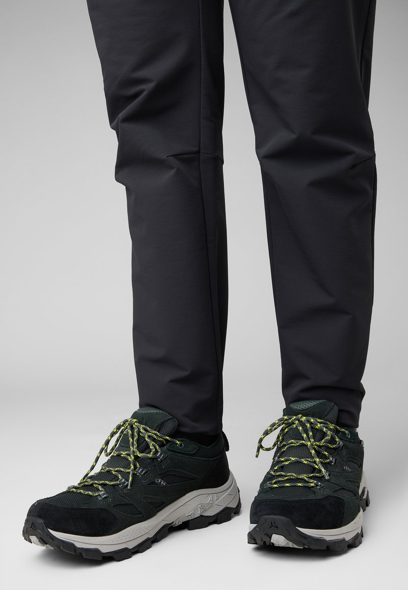Scarpe da esterno nere con materiale testurizzato e suola spessa, caratterizzate da lacci e dettagli verde brillante, abbinate a pantaloni scuri.