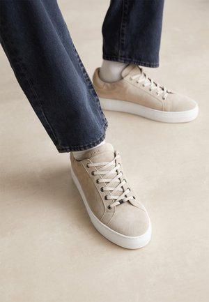 Osoba nosząca beżowe zamszowe trampki z białą podeszwą i białe skarpetki, zestawione z ciemnoniebieskimi dżinsami na jasnej podłodze.