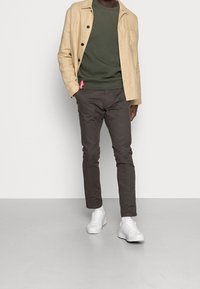 Lichtbeige linnen jasje over een donkergroen sweatshirt, gecombineerd met grijze broek en witte sneakers, met een rode label op de taillebaan.