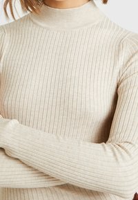 Personne portant un col roulé à manches longues côtelé beige, les bras croisés, mettant en valeur la texture tricotée et les détails du tissu.