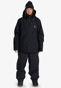 Svart vattentät jacka med justerbar huva och framfickor med dragkedja, kombinerad med lösa svarta byxor och mörka skor, tillverkad av hållbara material.