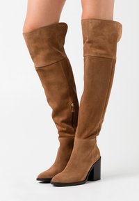 Bruine suede laarzen tot boven de knie met een spitse neus en een blokhak. Beschikt over een rits aan de binnenzijde en een omgevouwen bovenrand.