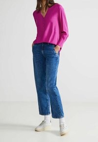 Blusa fucsia a maniche lunghe con scollo a V e dettagli con bottoni, abbinata a jeans a gamba dritta blu e sneakers bianche con accenti testurizzati.
