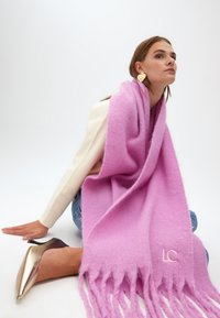Lyserød, blød uld tørklæde med frynser og broderet "LC" logo. Modellen bærer det draperet over en kremfarvet top og sidder i blå jeans.