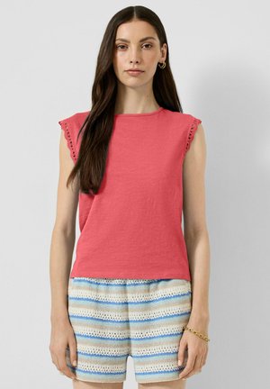 Kobieta o długich brązowych włosach nosi koralowy, bezrękawnik z koronkową lamówką oraz niebieskie, białe i beżowe, w paski, dziergane szorty, stojąc na jednolitym tle.