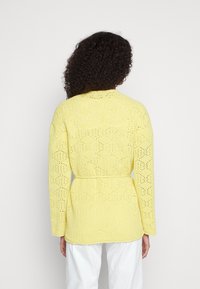 Żółty dziergany kardigan z wzorem w romby, długimi rękawami i paskiem wiązanym w talii, pokazany od tyłu na neutralnym tle.