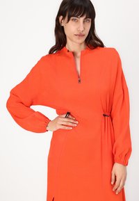 Felrood oranje jurk met een ritskraag, lange pofmouwen en verstelbare zijkoordjes. Glad materiaal met zijsplitten.