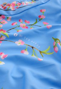 Tela azul con patrón floral rosa y hojas verdes, con texto "6 by Ted Baker" en letras blancas sobre una superficie ligeramente arrugada.