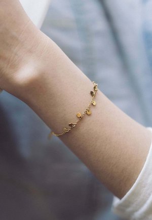 Bracelet en or avec de petits charms de forme irrégulière sur une chaîne délicate, porté à un poignet à la peau claire, affichant une texture lisse.