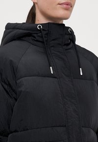 Svart puffjakke med teksturert finish, med hette, sølvfarget maskinvare og justerbare snorene. Framlommene er synlige.