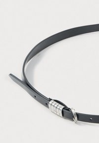 Melna ādas josta ar gludu tekstūru, kurai ir sudraba cilindriskā sprādze ar iegravētām burtiem un vairākiem regulēšanas caurumiem.