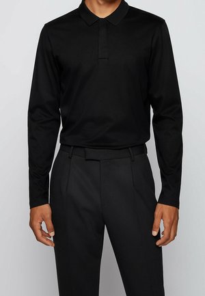 Man draagt een nauwsluitend zwart poloshirt met lange mouwen, ingestopt in een zwarte nette broek met riemlussen, tegen een effen lichte achtergrond.