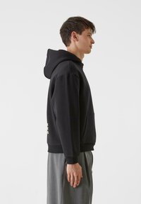 Črna kapuča z žepom za kenguru in rebrastimi manšetami. Model prikazan v profilu, v kombinaciji s sivimi hlačami. Minimalistična zasnova.