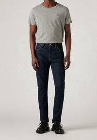 Camiseta gris de manga corta con un logo rojo, combinada con jeans de mezclilla oscuros y zapatos negros. Enfatiza un diseño casual, ajustado y con un detallado mínimo.