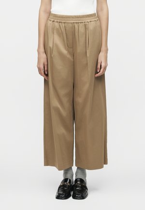 Person iført beige vidde bukser med elastisk talje, grå sokker og sorte slip-on loafers, stående mod en ensfarvet baggrund.