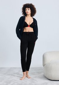Black hoodie with red accents, black bra with logo, and black joggers. Soft texture and fitted design. Barefoot on a light textured surface.