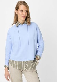 Felpa con cappuccio azzurro chiaro a maniche lunghe indossata sopra una camicia a righe verdi e bianche con colletto, abbinata a pantaloni color chiaro.