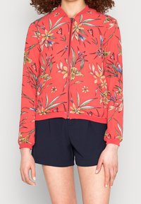 Blouson aviateur rouge avec imprimé floral en jaune, bleu et vert, fermeture éclair frontale, poignets et taille élastiqués. Associé à un short bleu marine.