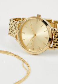 Gold-tone watch with a round face, minimalist markers, and a textured metal bracelet. Includes a gold chain beside the watch.