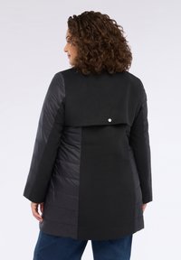 Chaqueta negra con una mezcla de tejido texturizado y paneles acolchados lisos. Presenta una solapa en la parte posterior y un sencillo detalle de botón metálico.