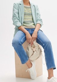 Personne portant un blazer vert clair, un pull en maille rayé, un jean bleu et des baskets blanches, assise sur un cube en bois et tenant un sac à main beige.