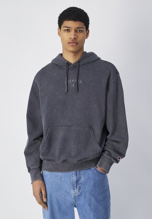 Sudadera gris oscuro de algodón con bolsillo tipo canguro, capucha con cordón y logo bordado en el pecho. Combinada con jeans de color azul claro.