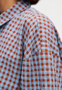 Camisa de manga corta confeccionada en tejido de algodón, con un patrón de cuadros azules y rojos, cuello clásico y detalles de costuras visibles.