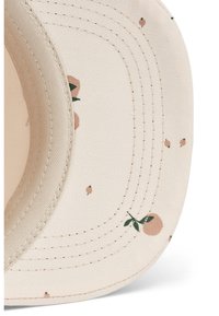 Bēša cepure ar ziedu rakstu, kurā redzami mazi persiku motīvi un zaļas lapas. Redzamas izšūšanas līnijas un izliekts saules bremzes malu.