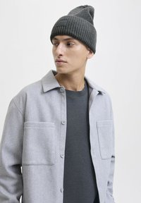 Jonge man in een donkergrijze gebreide beanie, antracietkleurige trui en lichtgrijze overhemdjas die naar de zijkant kijkt tegen een effen achtergrond.