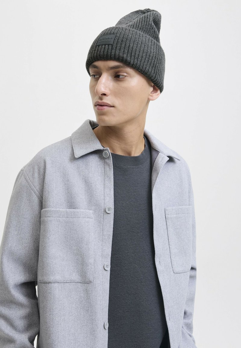 Jonge man in een donkergrijze gebreide beanie, antracietkleurige trui en lichtgrijze overhemdjas die naar de zijkant kijkt tegen een effen achtergrond.