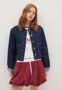 Jeune femme aux cheveux roux portant une veste en jean foncé, un t-shirt graphique blanc et un short à cordons bordeaux, se tenant contre un mur blanc uni.