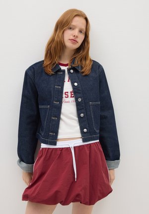 Young woman with red hair wears dark denim jacket, white graphic t-shirt, and maroon drawstring shorts, standing against a plain white wall.